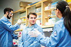 Photograph: Two lab technicians in blue safety coats talk in the foreground of a lab, while a coworker pipets in the background.