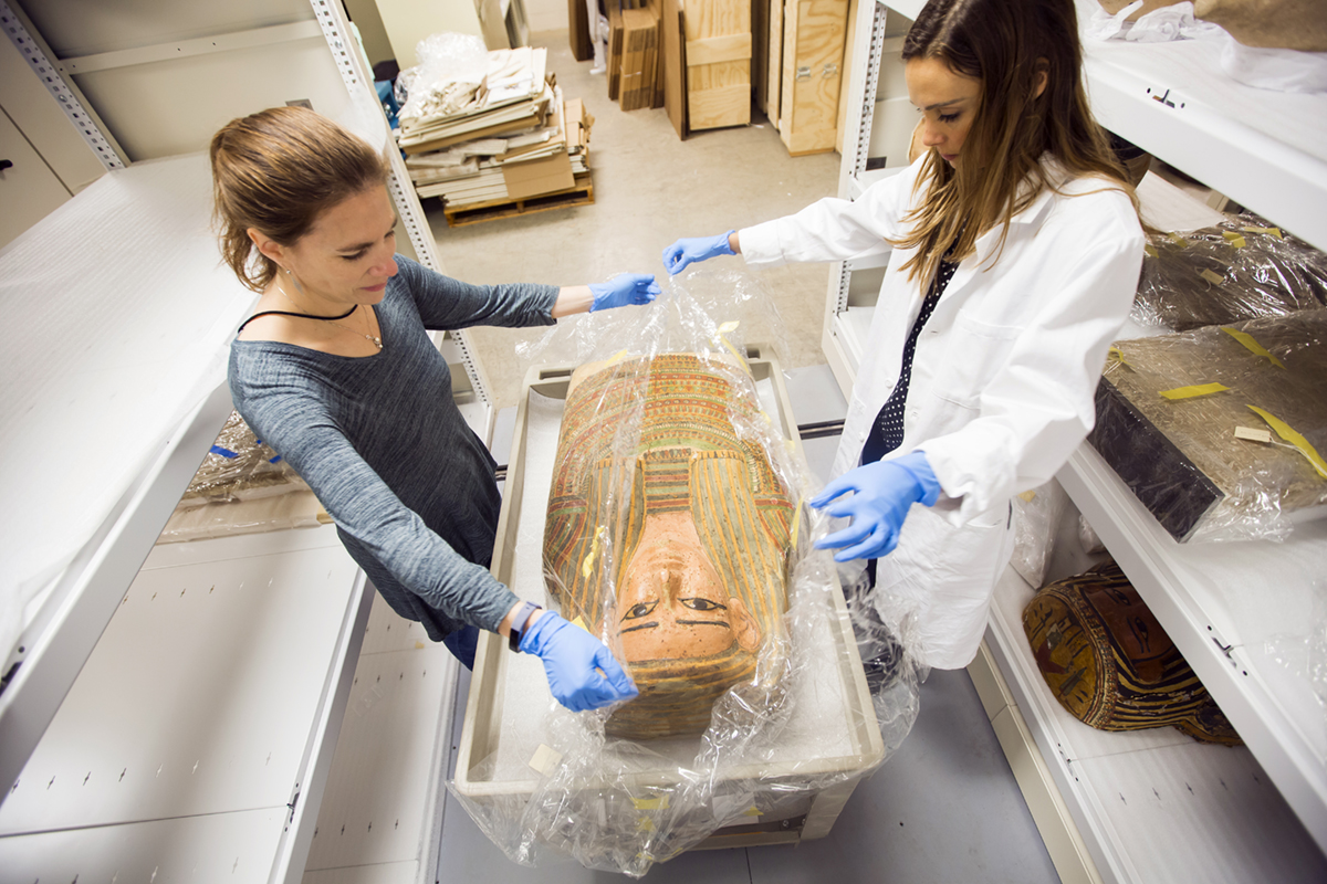 Two women, one in a white lab coat, both wearing blue safety gloves, very carefully unwrap an Egyptian artifact from clear plastic wrapper in which it has been shipped.