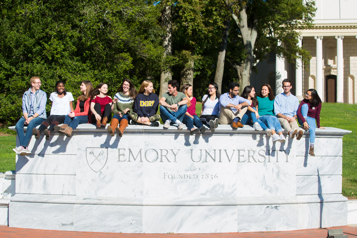 About 20 Emory University students sit atop a long marble wall at the gate to the university. The Emory university logo is carved on the wall below their feet.