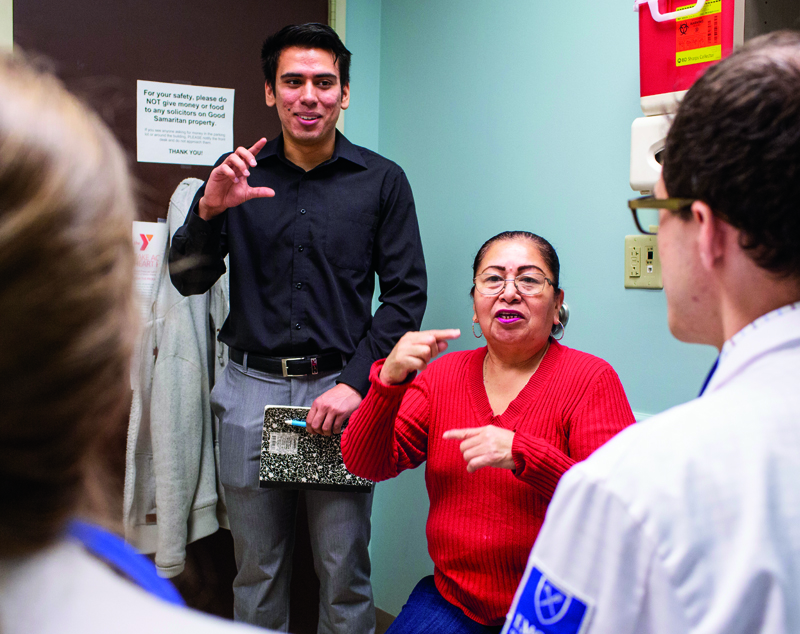 Student interpreter Aguilar laughs while gesturing, interpreting for a patient in a bright red sweater as Emory medical students in white coats look on.