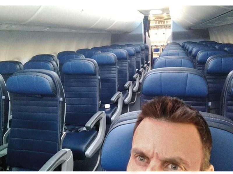 A selfie taken on a plane showing a man's face from the eyes up. Behind him are rows of empty, blue airplane seats.