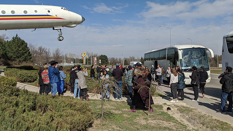 People congregate near buses at an airport as a plane lands.