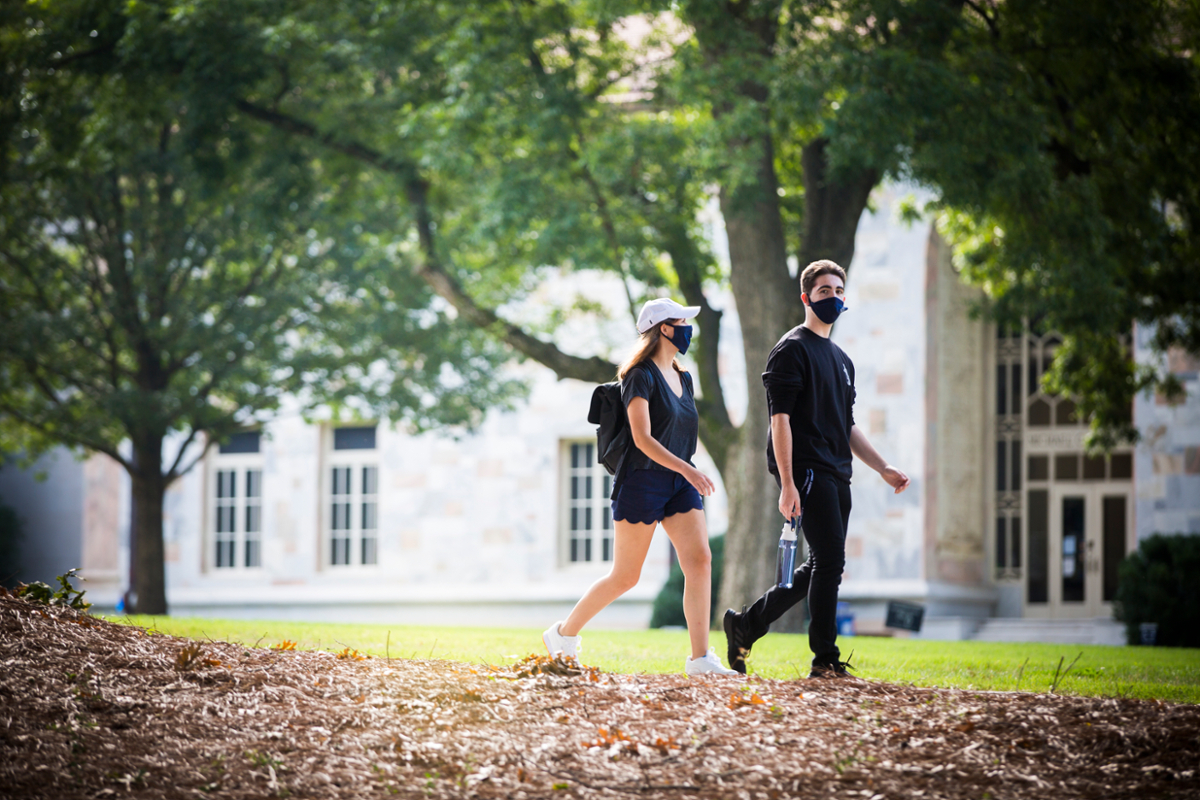 Two Emory students walk on the Emory Quadrangle, both wearing masks
