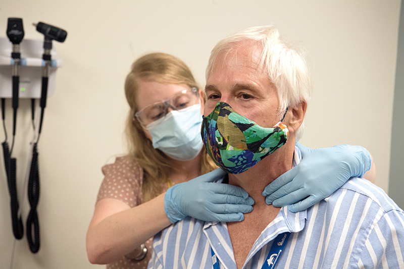 A nurse wearing blue latex gloves examines a man's neck from behind. Both are masked.