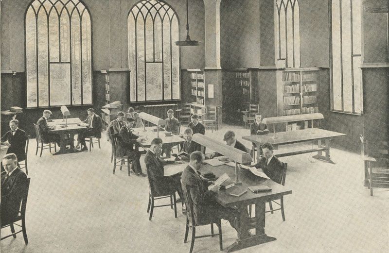 A black and white photo pictures several males wearing suits and ties seated at tables reading books and papers in the Theology Building Room