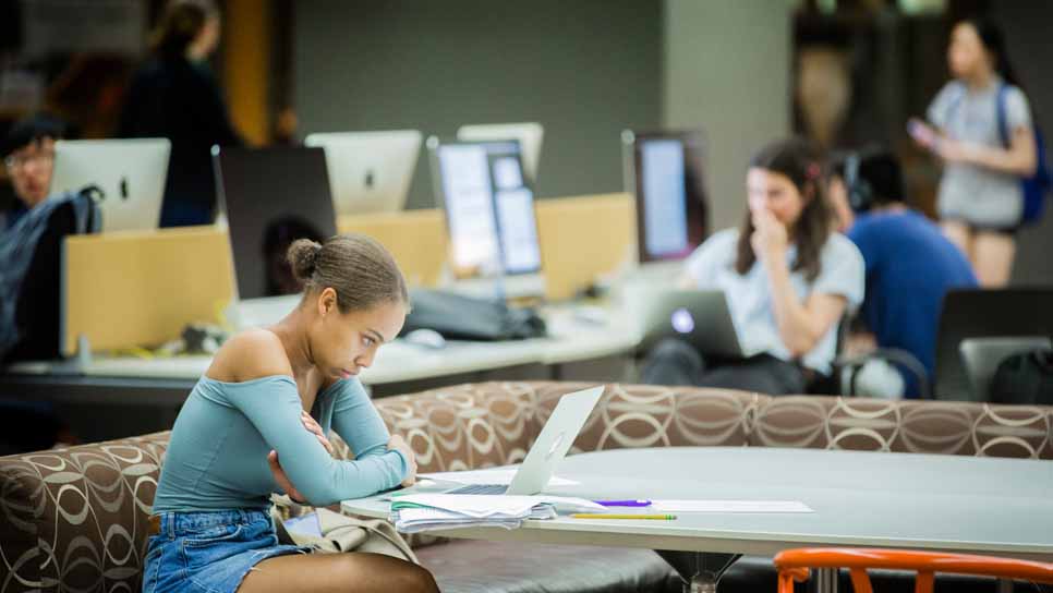 student in foreground in blue shirt sitting at table looking at her computer