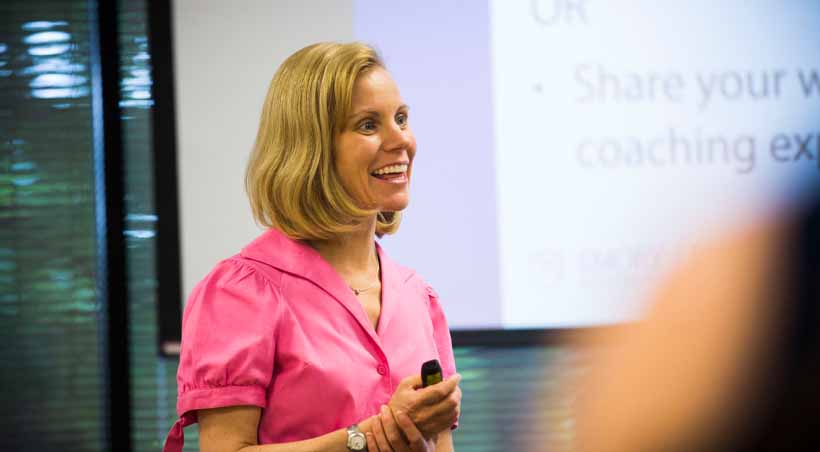 teacher in pink blouse stands in front of screen presenting