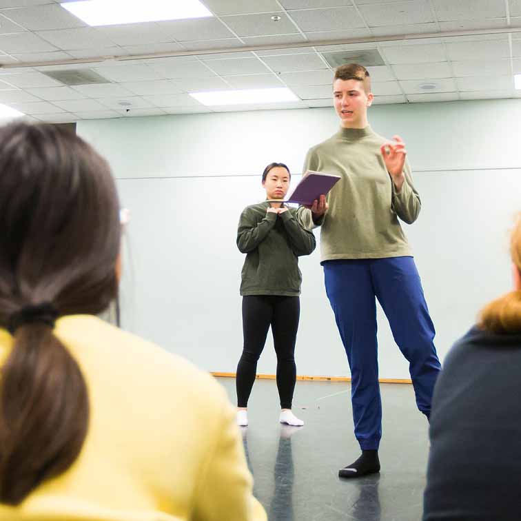 student standing on dance floor with book in hand, foot pointed