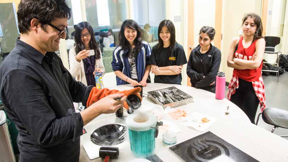 male professor conducting some type of experiment while students gather around the table to watch