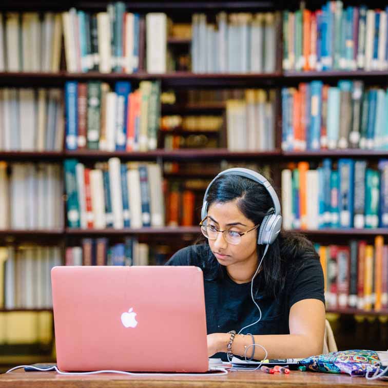 student at table with pink computer and headphones on in front of library stacks in background