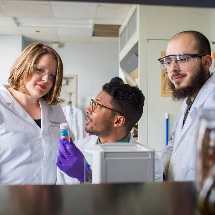 undergraduate student in purple gloves holds up vial for professor and graduate studenta