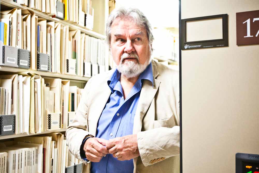 Collector Raymond Danowski in a rumpled linen jacket and bright blue shirt posed within library stacks 